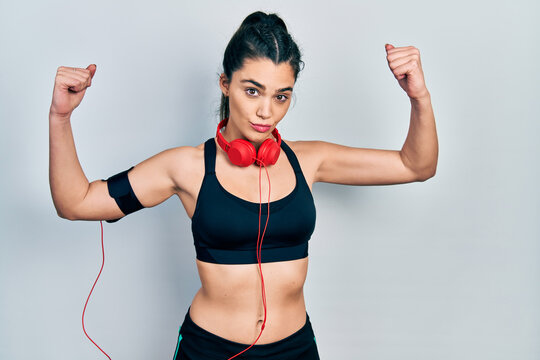 Young hispanic girl wearing gym clothes and using headphones showing arms muscles smiling proud. fitness concept.
