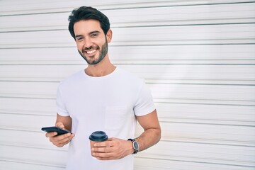 Fototapeta premium Young hispanic man smiling happy using smartphone and drinking take away coffee at city.