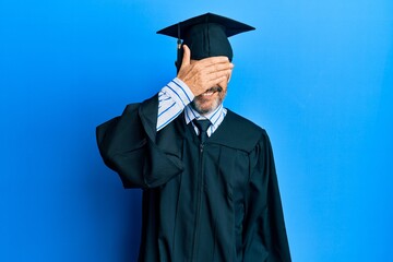 Middle age hispanic man wearing graduation cap and ceremony robe smiling and laughing with hand on face covering eyes for surprise. blind concept.