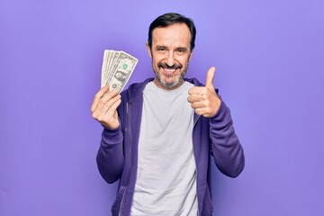 Middle age handsome man holding bunch of dollars banknotes over isolated purple background smiling happy and positive, thumb up doing excellent and approval sign