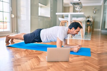 Middle age man with beard training and stretching doing exercise at home looking at sport video on...