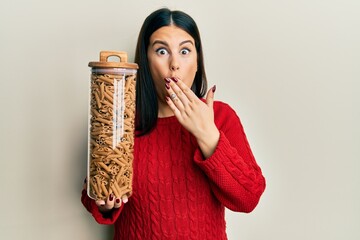 Beautiful brunette woman holding jar with macaroni pasta covering mouth with hand, shocked and...