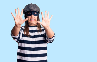 Young beautiful brunette woman wearing burglar mask afraid and terrified with fear expression stop gesture with hands, shouting in shock. panic concept.