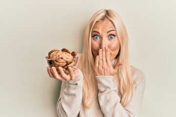 Young blonde woman holding bowl with chocolate chips cookies covering mouth with hand, shocked and...