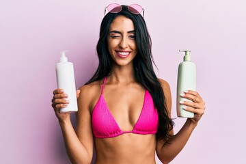 Beautiful hispanic woman wearing bikini holding sunscreen lotion winking looking at the camera with...