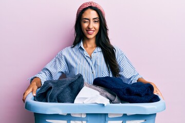 Beautiful hispanic woman holding laundry basket winking looking at the camera with sexy expression,...