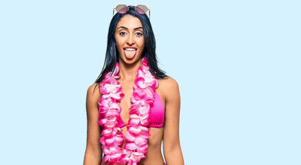 Beautiful hispanic woman wearing bikini and hawaiian lei sticking tongue out happy with funny expression. emotion concept.