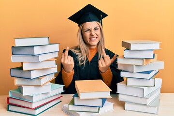 Young caucasian woman wearing graduation ceremony robe sitting on the table showing middle finger doing fuck you bad expression, provocation and rude attitude. screaming excited