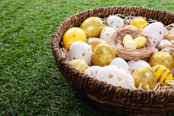 Many different Easter eggs in wicker basket on green grass, closeup
