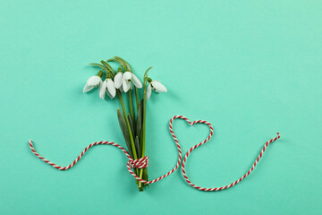 Beautiful snowdrops with traditional cord martisor on turquoise background, flat lay. Symbol of first spring day