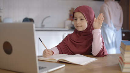 The little female student wearing a headscarf is watching the lessons on the internet with a...