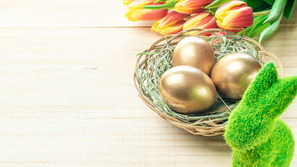 Easter eggs basket. Golden egg in basket with spring tulips, white feathers on wooden background in Happy Easter decoration. Traditional decoration in sun light.