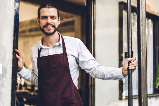 Portrait of handsome bearded barista man small business owner smiling outside the cafe or coffee shop.Male barista standing at cafe