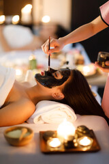 Masseurs hand do holding glass and brushing a facial mask herb black cream to on customer's face as...
