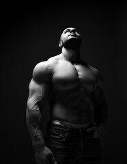 Fototapeta na wymiar Black and white portrait of strong muscular man, sportsman, athlete, fitness trainer with perfect built body stands shirtless and looks up over dark background