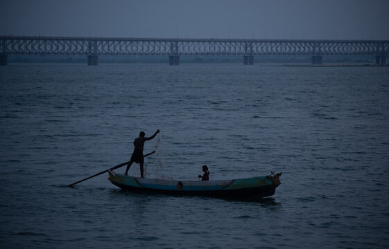 Rajahmundry, Andhra Pradesh, India, February,08, 2021 : A silhouetted fisherman adjusts his fishing net to cast in the river godavari in Rajamahendravaram near the Road cum Railway Bridge