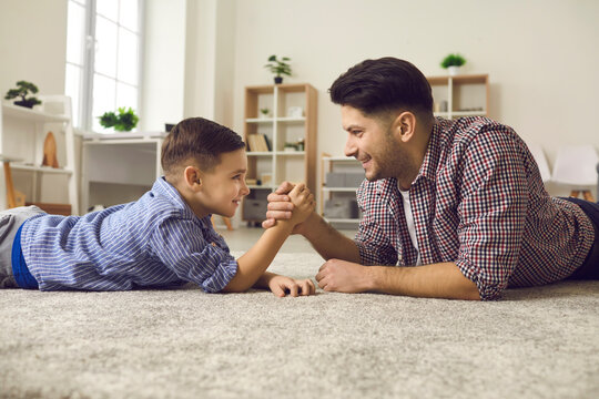 Handsome young father in casual clothes and his cute little son competing in arm wrestling while lying on floor. Father and son look into each other's eyes and smile. Family has fun together.