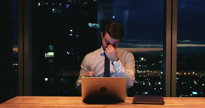 Stressed young caucasian businessman taking off eyeglasses, having painful feelings or dry eyes syndrome due to computer overwork, working overtime alone late in modern office, workaholic concept.