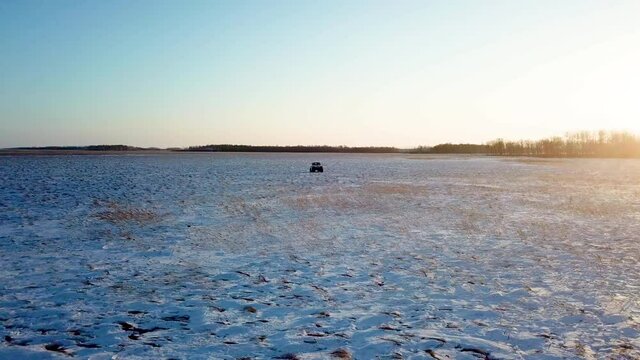 View from above. A light jeep on large marsh wheels rides across an endless snow field against the backdrop of the setting sun. The swamp vehicle travels across the Russian field in winter.
