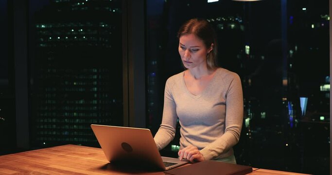 Focused young caucasian businesswoman employee working on computer, solving problems or making difficult decision, hardworking alone until late in modern office with panoramic night city view.
