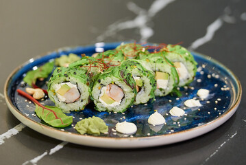 A close-up photo of sushi rolls with wakame seaweed, Philadelphia cheese, and shrimps on a blue ceramic plate with sauce, cashew, and wasabi. Black stone table.