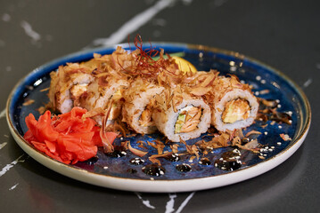 A close-up photo of fresh sushi rolls with tuna shavings, Philadelphia cheese, and eel on a blue ceramic plate with sauce, lobules of kumquat, and leaves. Black stone table.