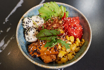 A top view of a poke bowl with tuna, avocado slices, wasabi, corn, peanut, sesame, and Philadelphia cheese on a black stone table.
