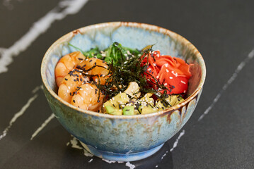 A close-up photo of a poke bowl with shrimps, Chuka Wakame seaweed, wasabi, avocado cubes, cashew, nori on a black stone table.