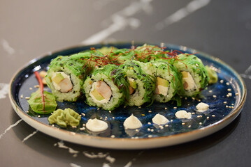 A close-up photo of sushi rolls with Chuka wakame seaweed, Philadelphia cheese, and shrimps on a blue ceramic plate with sauce, cashew, and wasabi on a black stone table.