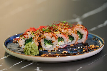 Sushi rolls with Chuka wakame seaweed, Philadelphia cheese, and shrimps on a blue ceramic plate with sauce, peanuts, ginger, and wasabi. Black stone table.
