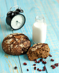 Dark rye bread loaf with crispy crust, glass of milk and alarm clock. Freshly bakery food in working day morning. Light rustic breakfast on light blue wooden table. Vertical 4x5 format. Soft focus.