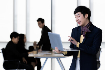 Portrait of LGBTQ transgender man office employee in casual suit standing using labtop notebook computer with elegant . and happy gesture with team colleagues blur in background