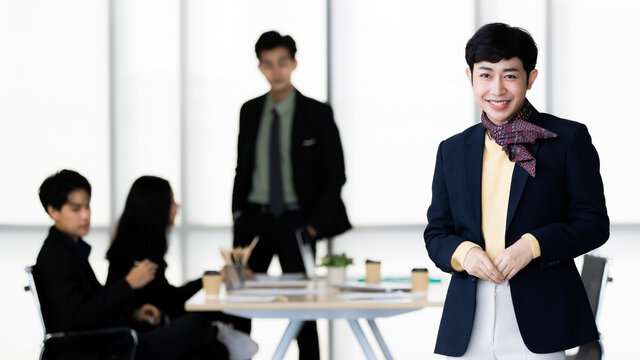 Portrait Of LGBTQ Transgender Man Office Employee In Casual Suit Standing And Pose To Camera With Cheerful And Happy Gesture With Team Colleagues Blur In Background.