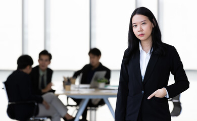 Young Asian businesswoman in black suit standing and posing to camera in office with her team in blur background