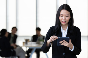 Young Asian businesswoman in black suit standing and holding tablet  computer posing to camera in office with her team talking in blur background