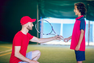 Male tennis coach in red clothes teacjing a boy to hold the racket