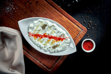 Tzatziki, cacık, or tarator is a traditional Turkish and Greek appetizer made from fresh yoghurt, cucumbers, spices and olive oil in a white plate. Dark background