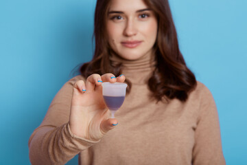 Beautiful European woman holding menstrual cup with pleasure, stands with flexible menstrual cup to insert in vagina during periods, protects from blood leakage, isolated on blue background.