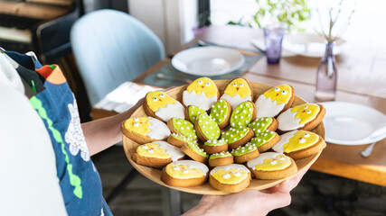 Festive table setting. A woman in an apron carries a platter of glazed Easter cookies in the shape of cute chicken-in-shell. Family dinner with sweets for children.