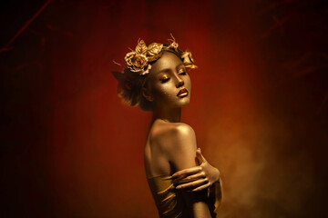 Fantasy portrait of woman with golden skin. Girl goddess in wreath, gold roses accessories. Beautiful face, steel glitter makeup. Artistic photo dark red background. Elf fairy princess. Fashion model