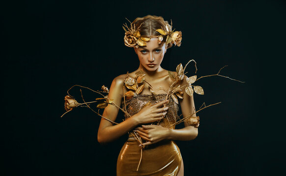 Beauty fantasy woman, face in gold paint. Golden shiny skin. Fashion model girl, image goddess. Glamorous crown, wreath roses, jewellery accessories. Professional metallic makeup. hand holds a branch.