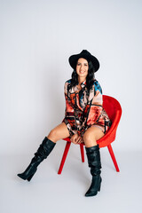 Beautiful young woman with colored dress and black hat sitting on a red chair white background self-confident
