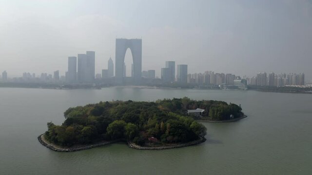 Aerial panning shot of green island at Jinji Lake near city against sky, drone flying near modern buildings - Suzhou, China