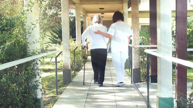 Back view of asian female doctor helps elderly woman with walker in the hospital garden patio - Slow motion