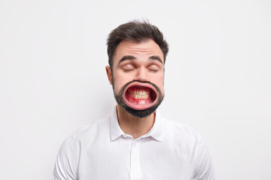 Portrait of unshaven European man keeps eyes closed opens mouth clenches teeth dressed in casual shirt isolated over white background. Funny guy makes grimace at camera. Face expressions concept