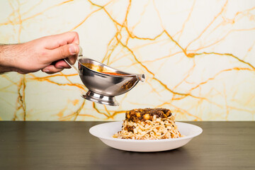 Famous traditional Arabic, Egyptian dish - Koshari. National Kushari in a white plate on dark wooden background with hand dropping tomato sauce