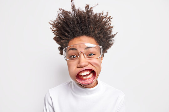 Close up shot of funny young Afro American woman with curly hair standing up has wide opened mouth wears transparent glasses casual turtleneck poses indoor exclaims loudly. Flight of imagination