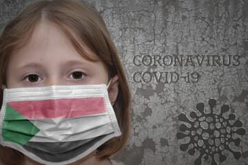 Little girl in medical mask with flag of sudan stands near the old vintage wall with text...