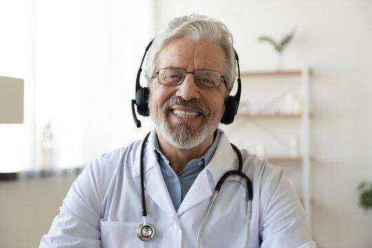 Headshot portrait of smiling mature Caucasian male doctor in headphones talk on video call in hospital. Profile picture of happy senior man GP or therapist in earphones consult patient on webcam.