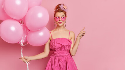 Obraz na płótnie Canvas Dreamy fashionable woman wears sunglasses and festive dress points away on blank space holds bunch of inflated balloons enjoys holiday isolated over pink background. Special occasion concept